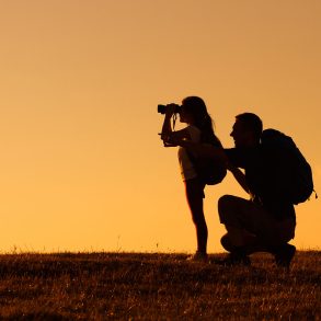 Silhouette of father and daughter with backpacks as daughter looks through binoculars