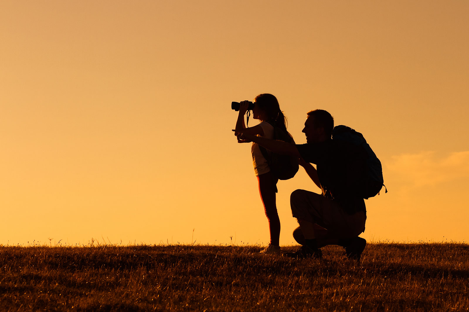 Silhouette of father and daughter with backpacks as daughter looks through binoculars